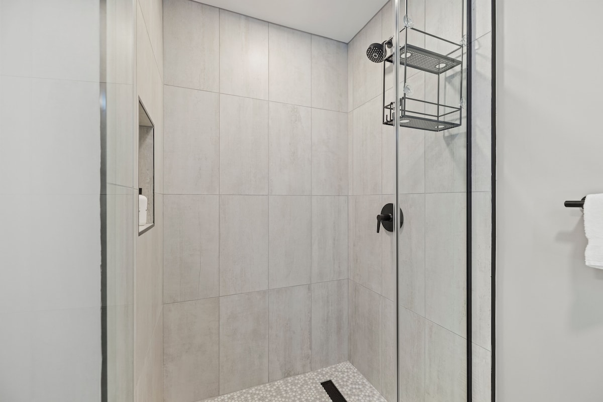 A modern shower with a glass door, a metal shelf, and a tiled floor.