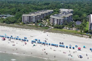 A beachfront hotel with multiple buildings, a sandy beach with umbrellas, and a forested area in the background. - BEAUTIFULLY RENOVATED, OCEAN VIEW - VILLAMARE. PICKLEBALL-TENNIS INCLUDED.   (Hilton Head Island)
