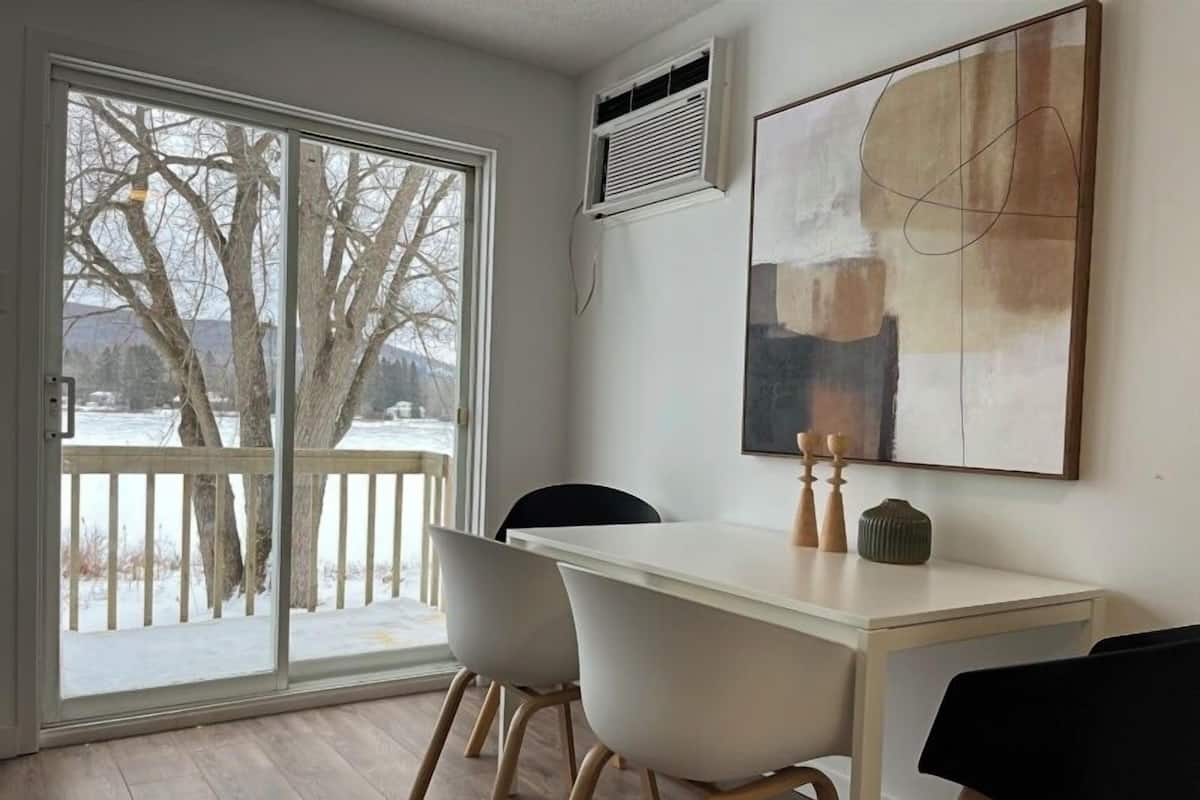 Een eethoek met een witte tafel en stoelen, een groot abstract schilderij aan de muur, en zicht op bomen en sneeuw buiten door een schuifdeur van glas.