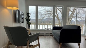 A modern living room with a grey armchair, a black sofa, and a floor lamp. A TV is mounted on the wall, and a plant is on a side table. - Hôtel Matha (Saint-Jean-de-Matha)