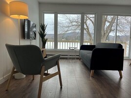 Un salón moderno con un sillón gris, un sofá negro y una lámpara de pie. Un televisor está montado en la pared, y hay una planta sobre una mesita auxiliar.