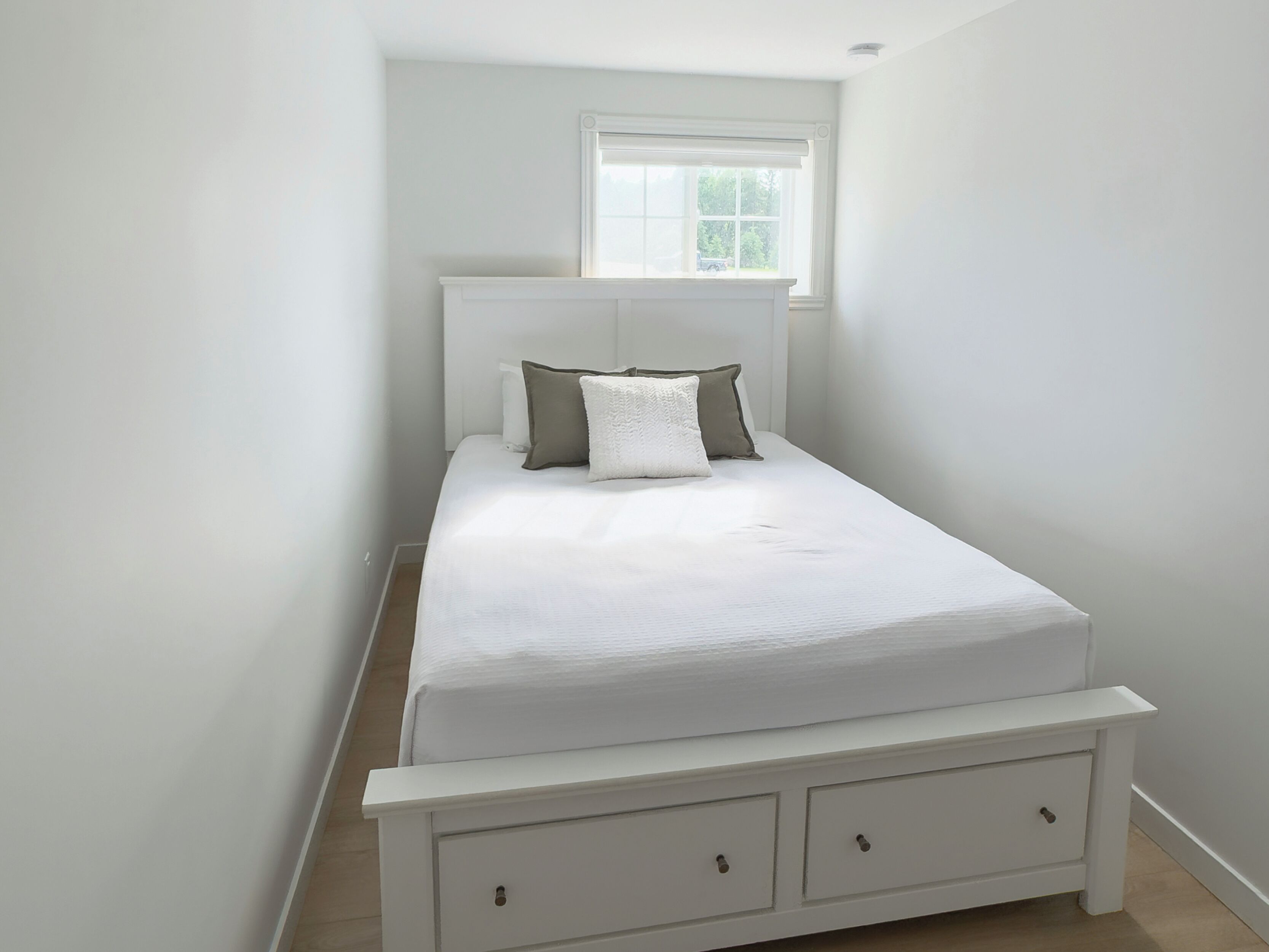 Uma cama pequena e bem arrumada, com cabeceira e peseira brancas, uma janela com vista para árvores e uma simples mesa de cabeceira com gavetas.