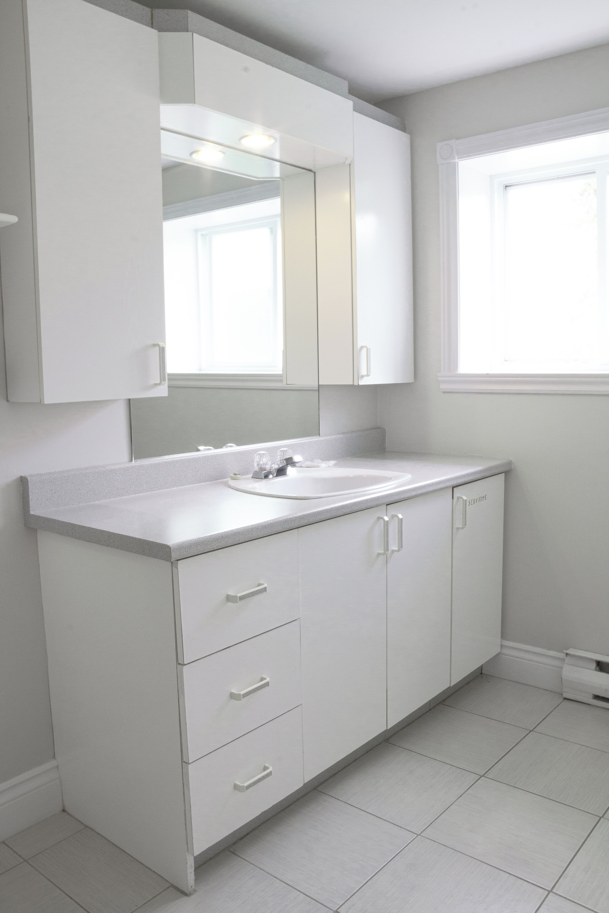 A modern bathroom with a large mirror, white cabinetry, and a window providing natural light.