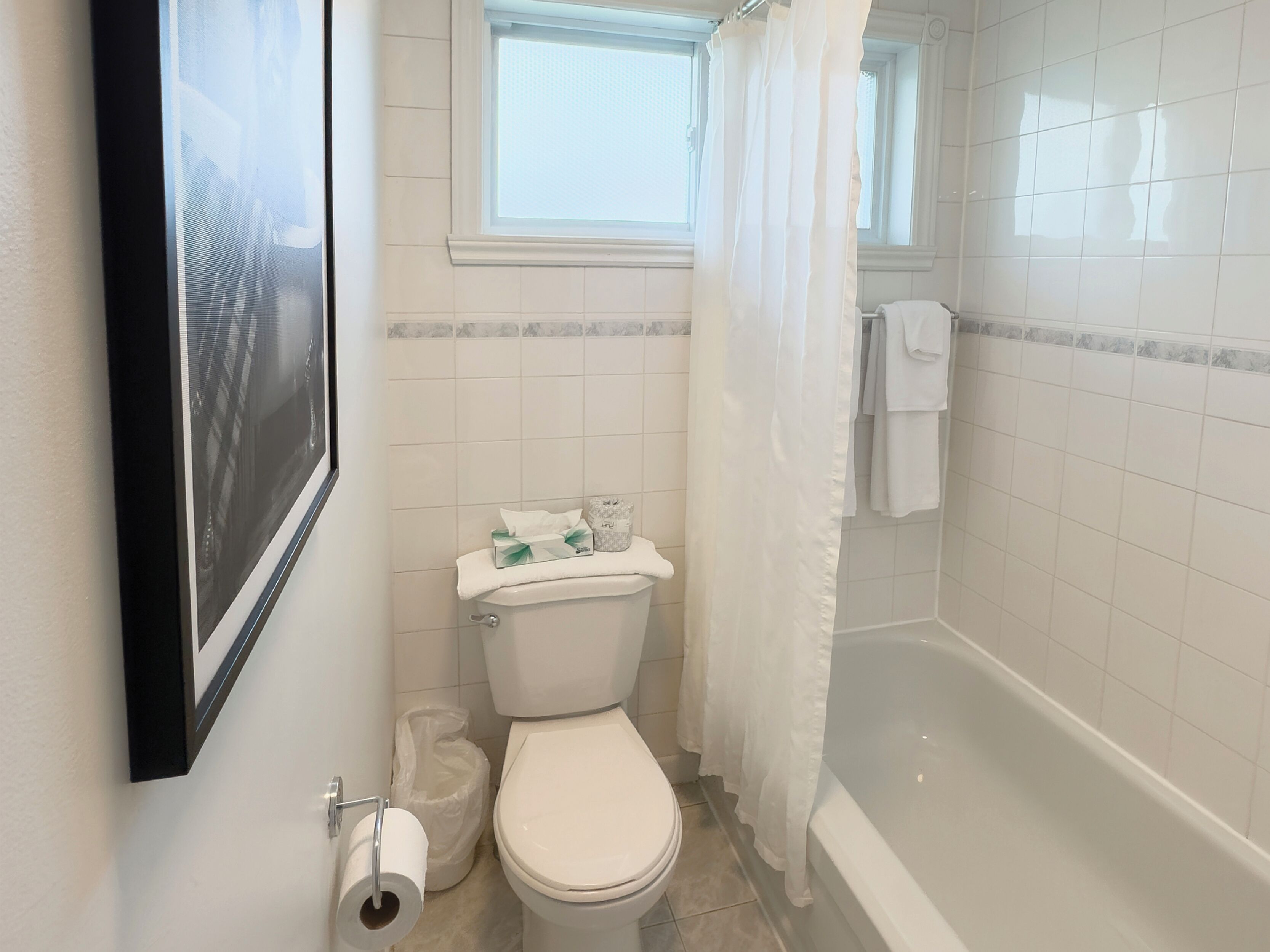 A bathroom with a toilet, a shower with a curtain, a window, and a framed picture on the wall.