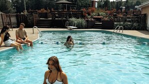 A swimming pool with clear water, a metal ladder, and a wooden deck area with chairs and umbrellas. - Hôtel Matha (Saint-Jean-de-Matha)