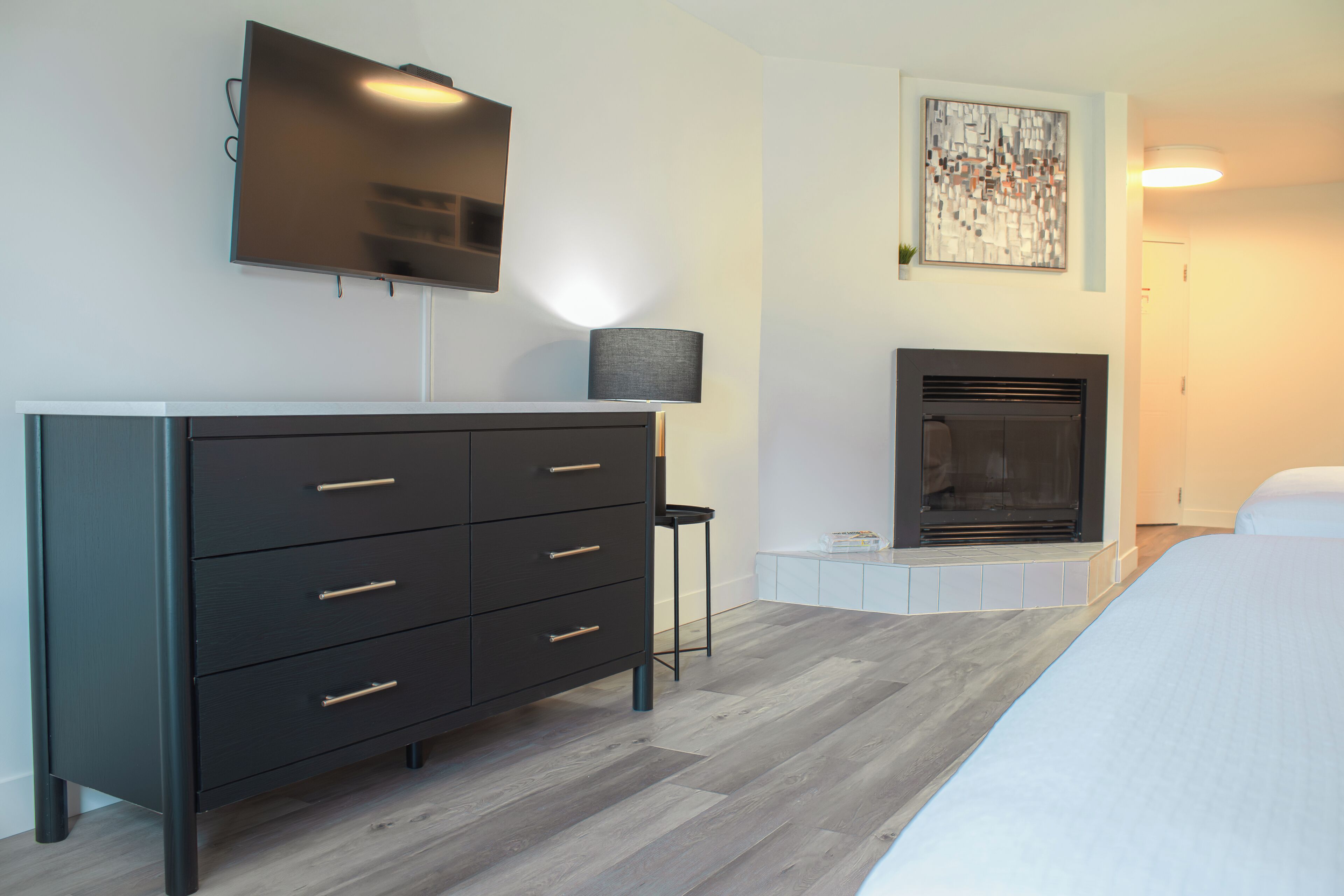 A modern bedroom with a large bed, a dresser, a flat-screen TV, a fireplace, and a small table.