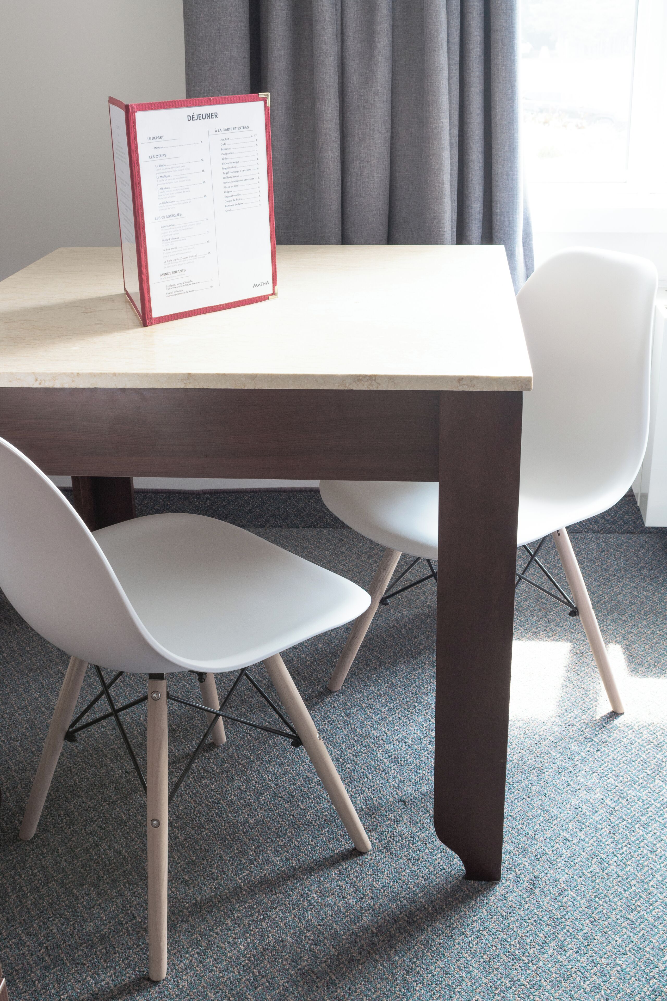 A dining table with a menu, two white chairs, and a window with curtains.