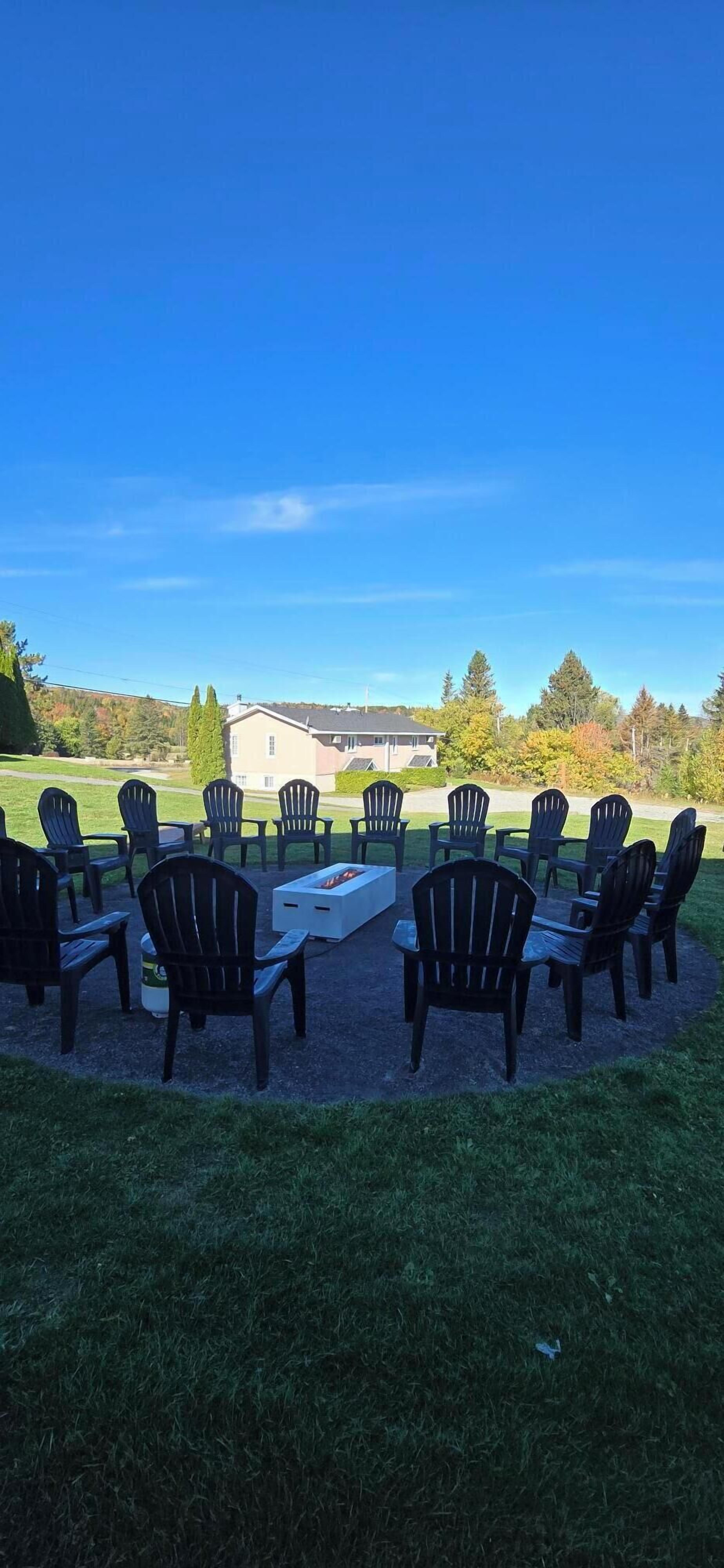 Uma área de estar externa com cadeiras Adirondack e uma lareira, cercada por um gramado e um edifício ao fundo.