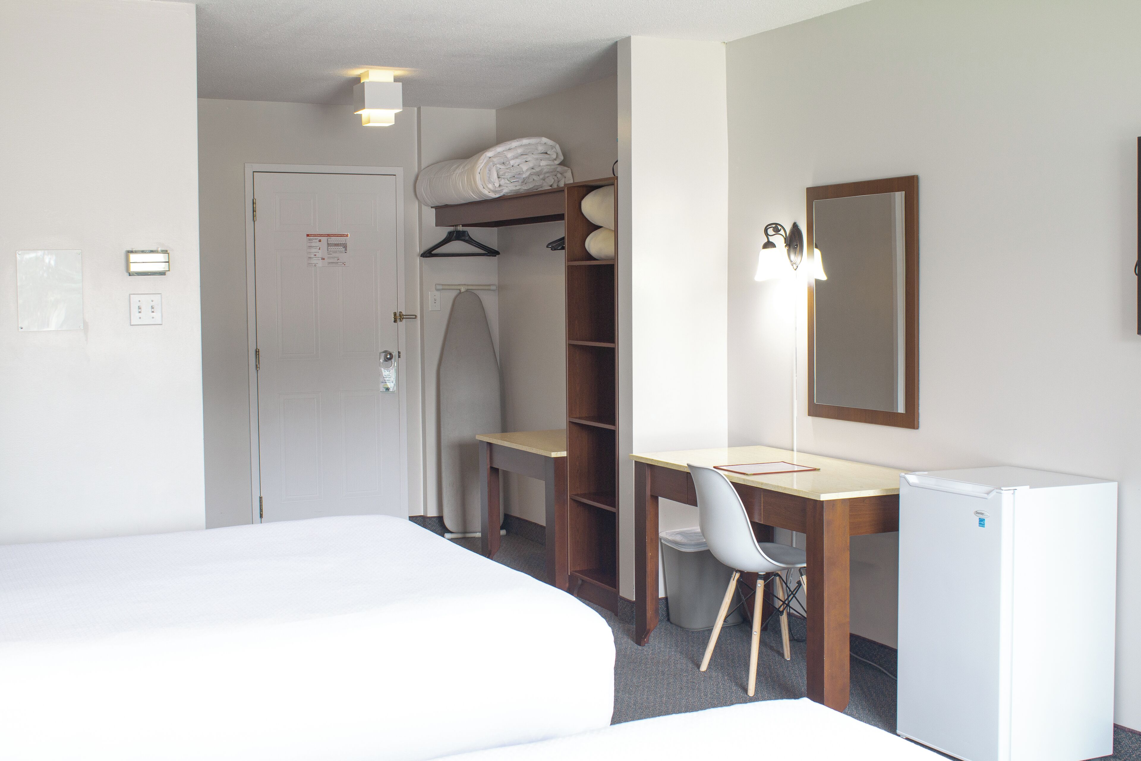 A hotel room with a bed, a desk, a chair, a wardrobe, and a small refrigerator.