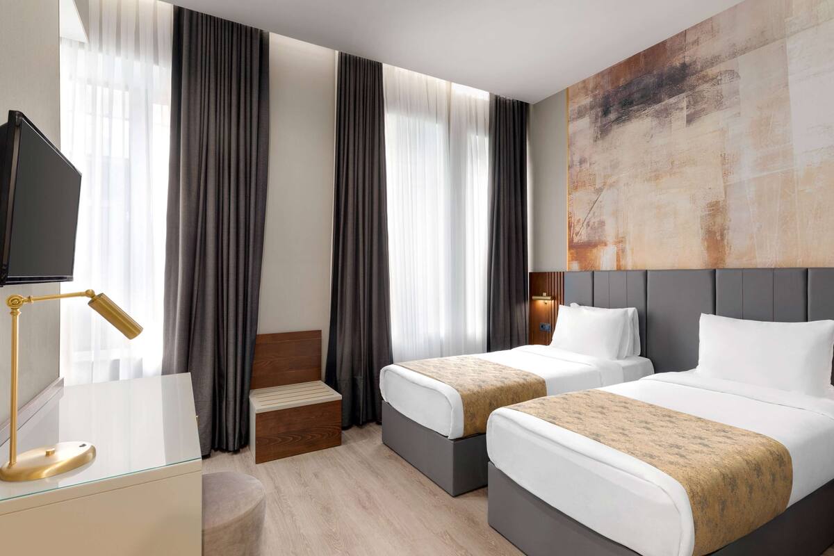 A hotel room with two beds, a desk with a lamp, a large abstract painting on the wall, and a staircase.