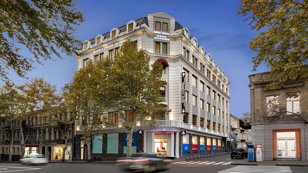 Un hôtel à plusieurs étages, de style architectural classique, qui se distingue par son entrée imposante et un grand arbre qui se trouve devant.