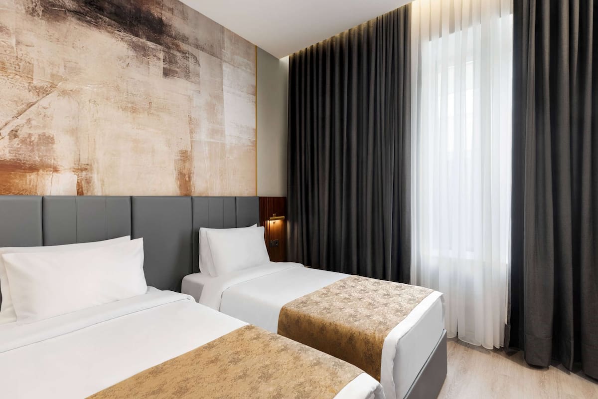 A hotel room with two beds, a large abstract painting on the wall, a window with dark curtains, and a wooden floor.