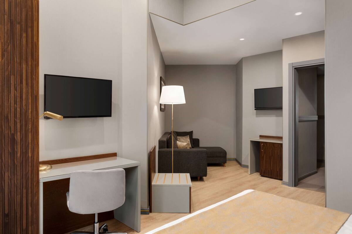 A modern hotel room with a flat-screen TV mounted on the wall, a desk with a chair, a sofa, and a floor lamp.