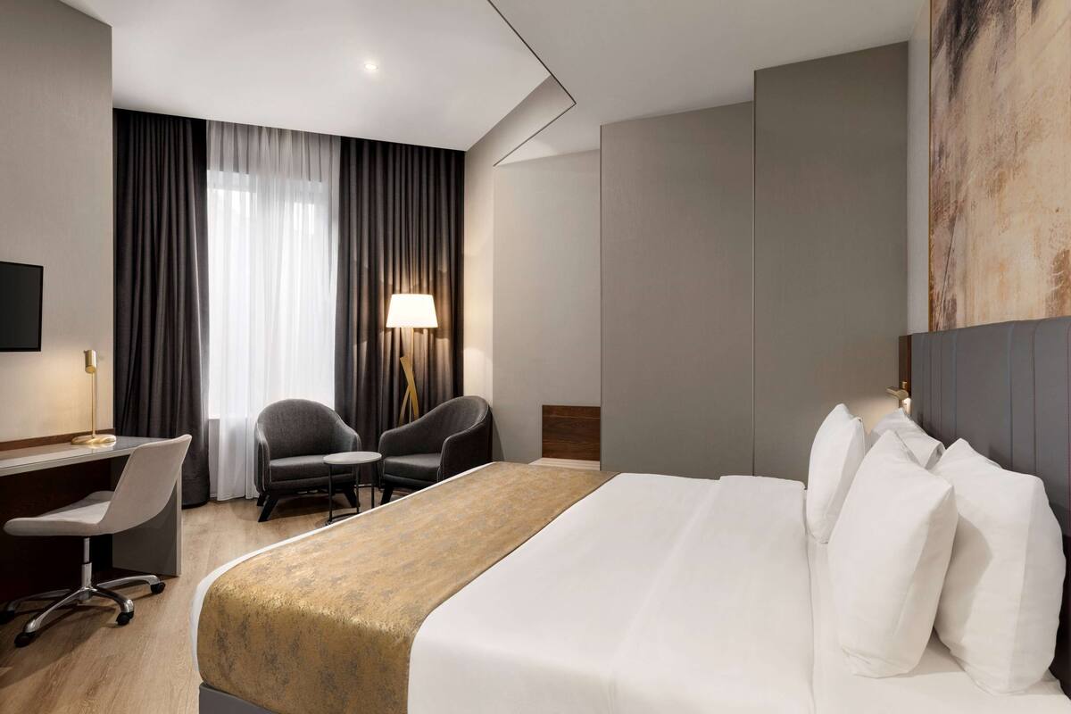 A hotel room with a large bed, a desk with a chair, a lamp, and a view of the outdoors through sheer curtains.