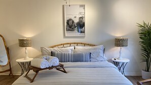 A neatly made bed with a striped bedspread, two bedside tables with lamps, a framed photo on the wall, and a plant on the right.