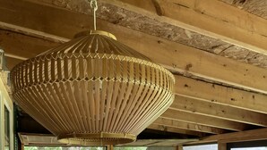Una habitación acogedora con una lámpara de techo grande de fibras naturales, una silla de madera con cojín estampado, una mesita con lámpara antigua y vistas a zona boscosa a través de amplios ventanales.