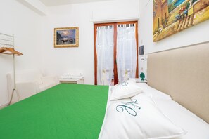 A hotel room with a green bedspread, a white headboard, a white sofa, a painting on the wall, and a door with curtains. - Villa Cristina (Piano di Sorrento)
