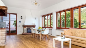 A spacious living room with a fireplace, wooden flooring, a sofa, and large windows offering a view of greenery. - Villa Cristina (Piano di Sorrento)
