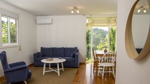 Une pièce avec un canapé bleu, une table blanche et une chaise en bois. On y trouve une grande fenêtre avec des rideaux, un luminaire au plafond et un appareil de climatisation.