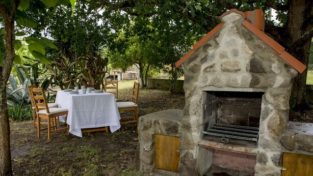 Un espace de restauration en plein air doté d’un four-barbecue en pierre, entouré de verdure et agrémenté d’une table dressée pour deux personnes.