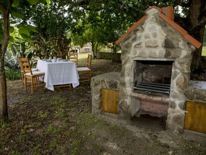 Een buiten eetgedeelte met een barbecueoven van steen, omringd door groen en een tafel gedekt voor twee personen.