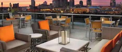 Terraço no telhado com assentos e mesas, com vista para o horizonte urbano ao entardecer.