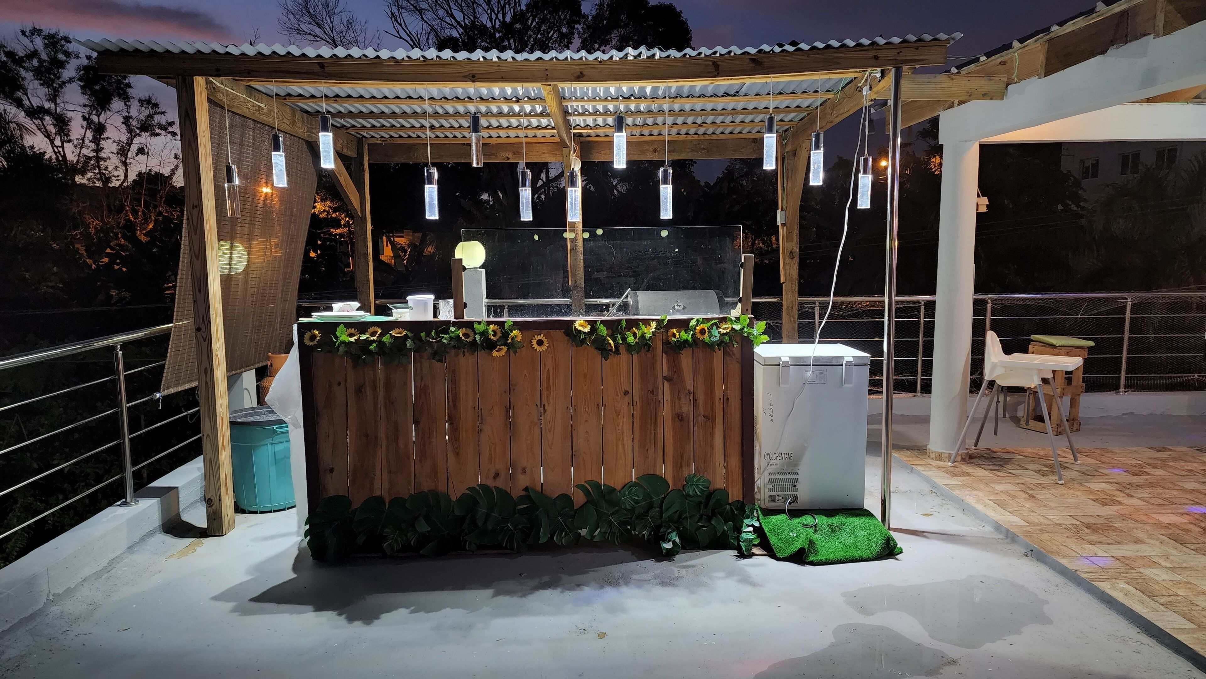 Area bar all'aperto con un bancone in legno, luci pendenti e una griglia.
