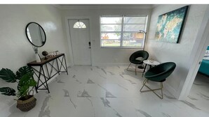 Une pièce avec une porte blanche, une fenêtre avec des stores, un miroir rond, une petite table sur laquelle se trouve un globe terrestre, deux chaises vertes et une carte du monde accrochée au mur.