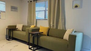Un salon avec un canapé vert, deux tables d’appoint et une fenêtre avec des rideaux.