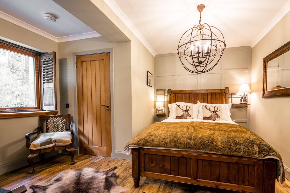 A bedroom with a wooden bed, a fur rug, a wooden chair, a window with blinds, a wooden door, and a chandelier.