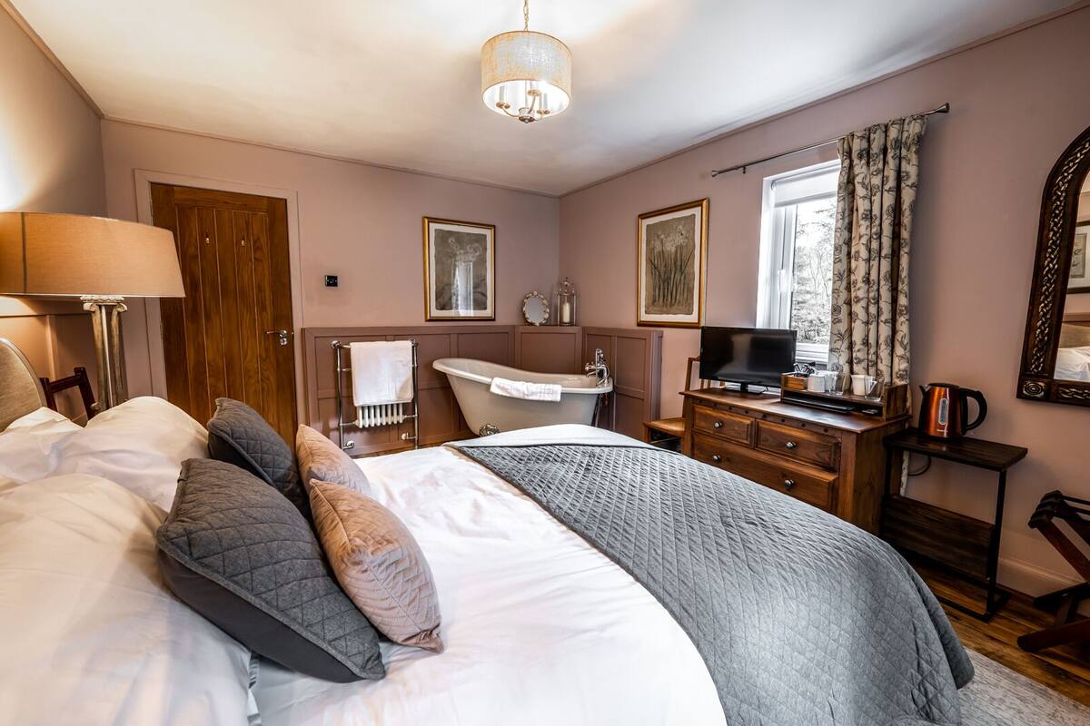A bedroom with a large bed, a bedside table, a dresser, a vanity with a mirror, and a window with curtains.