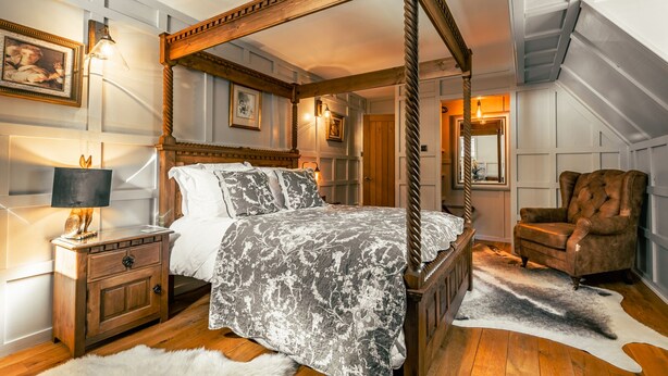 A four-poster bed with a patterned bedspread, a wooden nightstand with a lamp, a chair, and a fur rug on a wooden floor. - White Rose Tower (Invergordon)