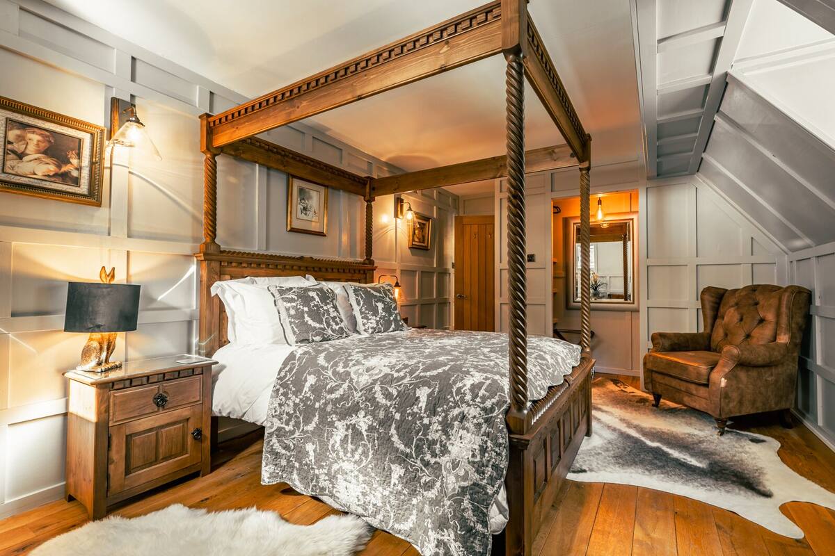 A four-poster bed with a patterned bedspread, a wooden nightstand with a lamp, a chair, and a fur rug on a wooden floor.