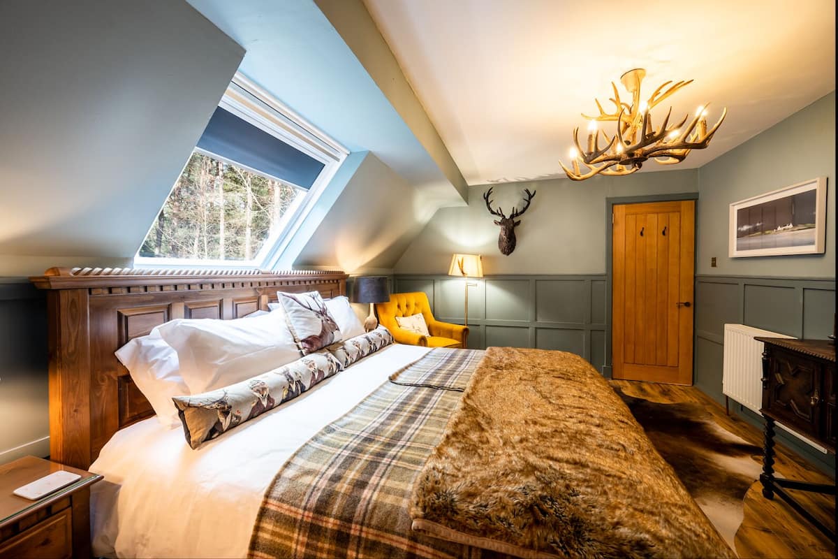 A bedroom with a sloped ceiling, a large bed, a chandelier, a deer head decoration, and a window with a view of trees.