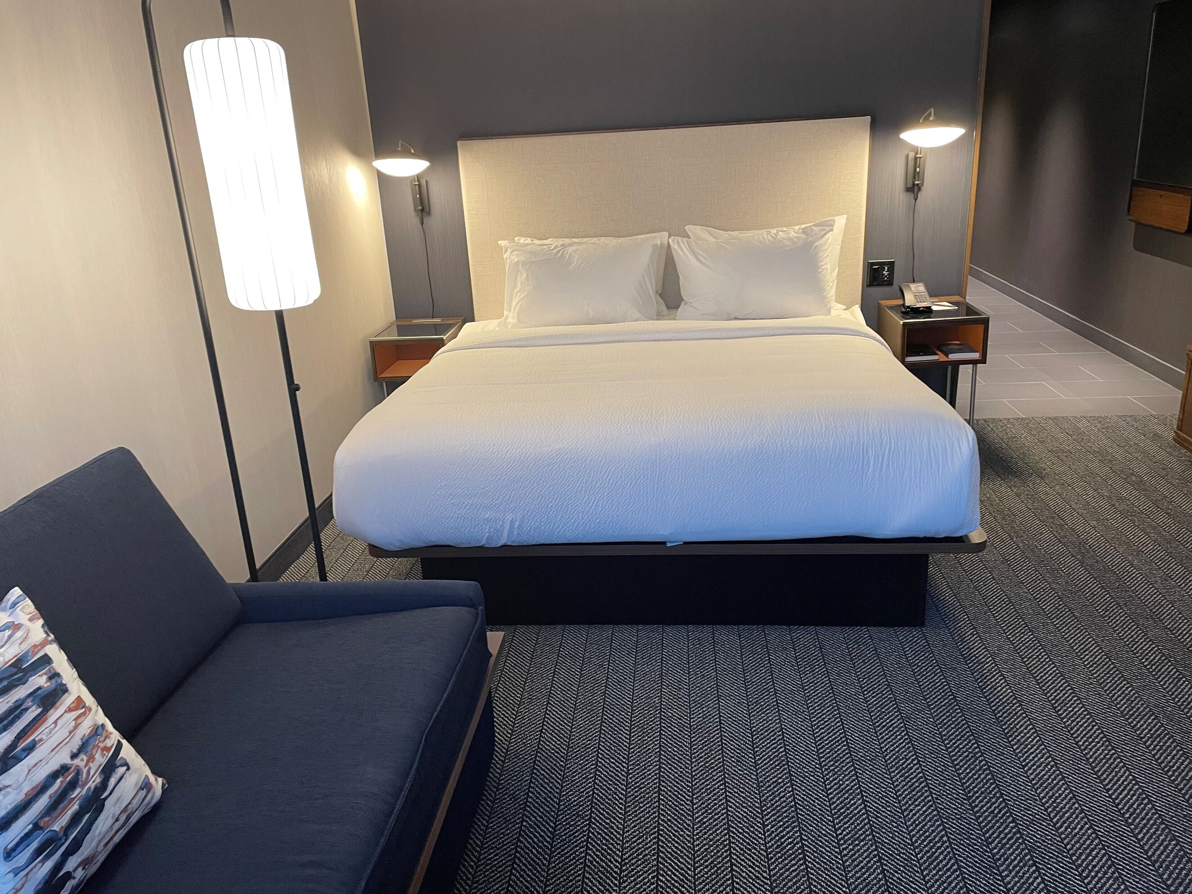 Quarto de hotel com uma cama grande, duas mesas de cabeceira, um criado-mudo, um sofá e um abajur.