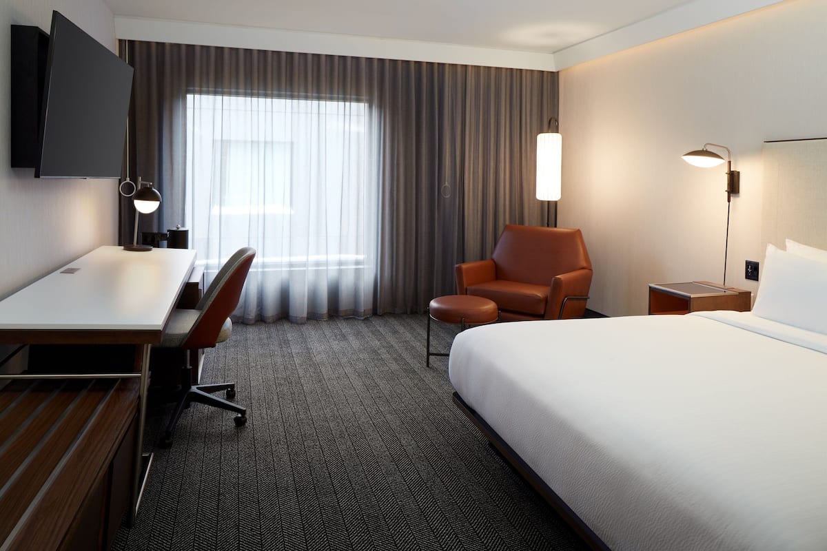 Ein Hotelzimmer mit Bett, Schreibtisch, Stuhl und einem an der Wand befestigten Fernseher.