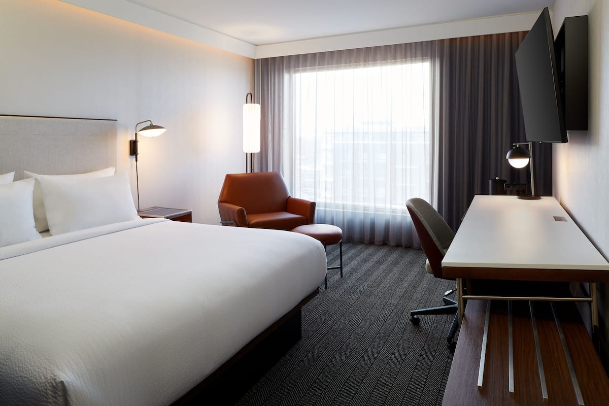 Ein Hotelzimmer mit einem großen Bett, einem Schreibtisch, einem Stuhl, einem Fernseher und einem Fenster mit Vorhängen.