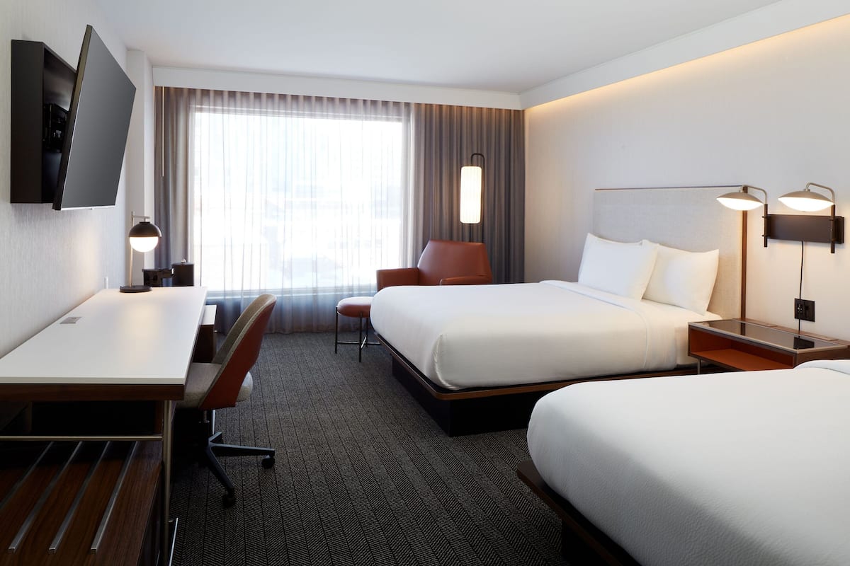 Ein Hotelzimmer mit zwei Betten, einem Schreibtisch, einem Stuhl, einem Fernseher und einem Fenster mit Vorhängen.