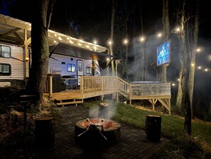 A campsite with a covered dining area, a fire pit, and a TV screen mounted on a tree.