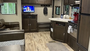 A compact RV kitchen with wooden cabinets, a built-in stove, a sink, and a television displaying a news broadcast. - Posh Mountain Camping w/ 120" Screen Theater ·Hot-Tub · Firepit · Hiking Trails (Jonesville)