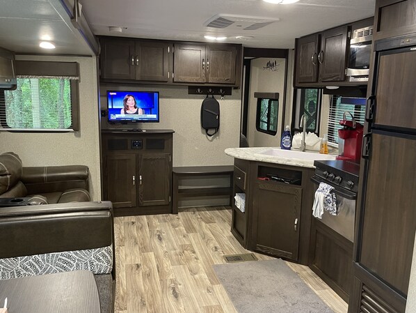 A compact RV kitchen with wooden cabinets, a built-in stove, a sink, and a television displaying a news broadcast.