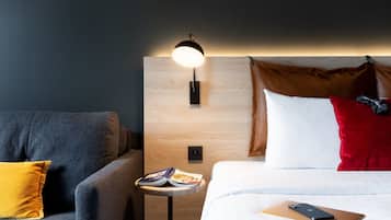 Een moderne hotelkamer met een houten hoofdeinde, een grijze bank, een klein tafeltje met tijdschriften en een bed met een laptop en een rode kussen.