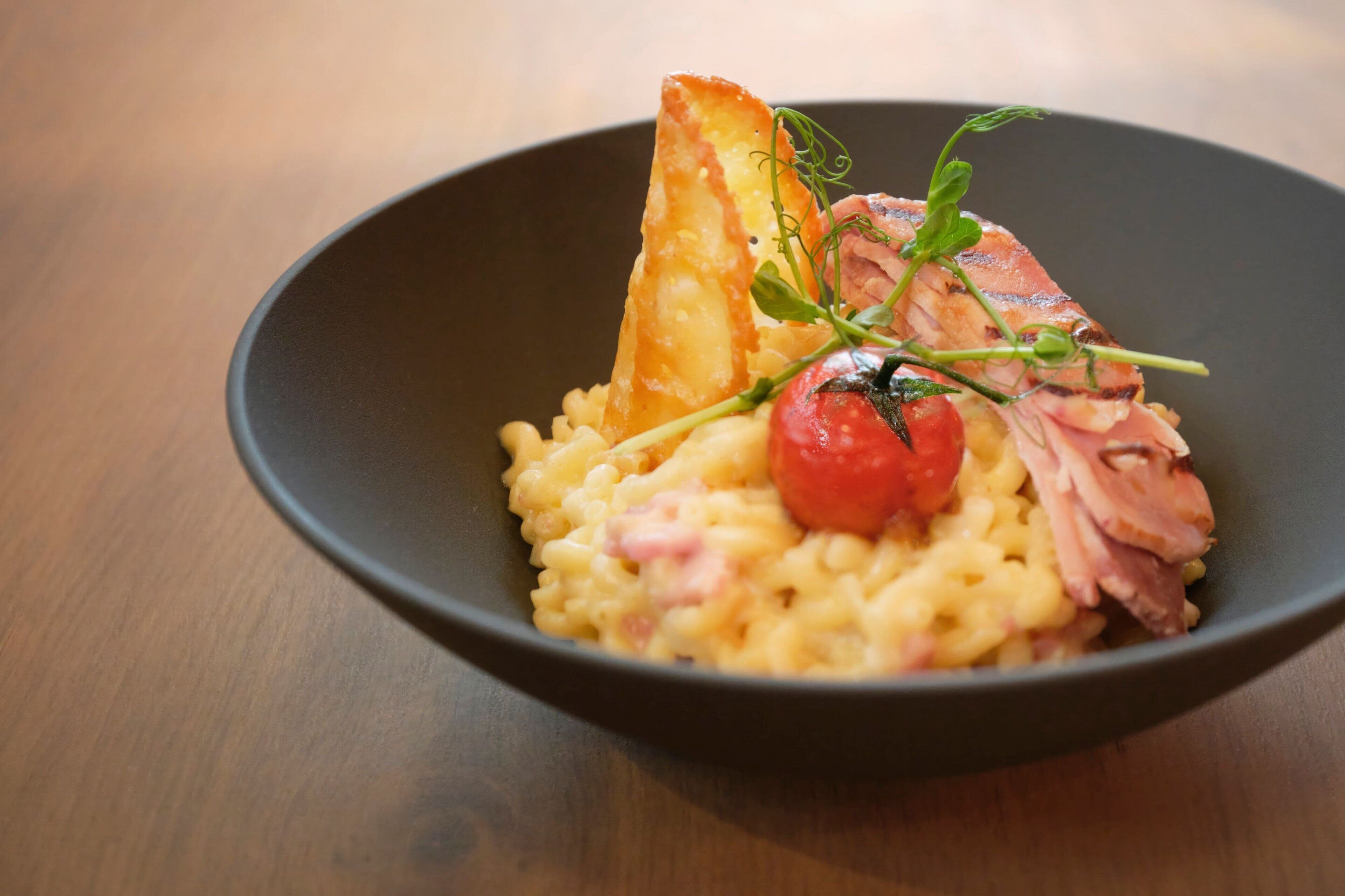 A bowl of food with pasta, a slice of meat, a cherry tomato, and a piece of bread.