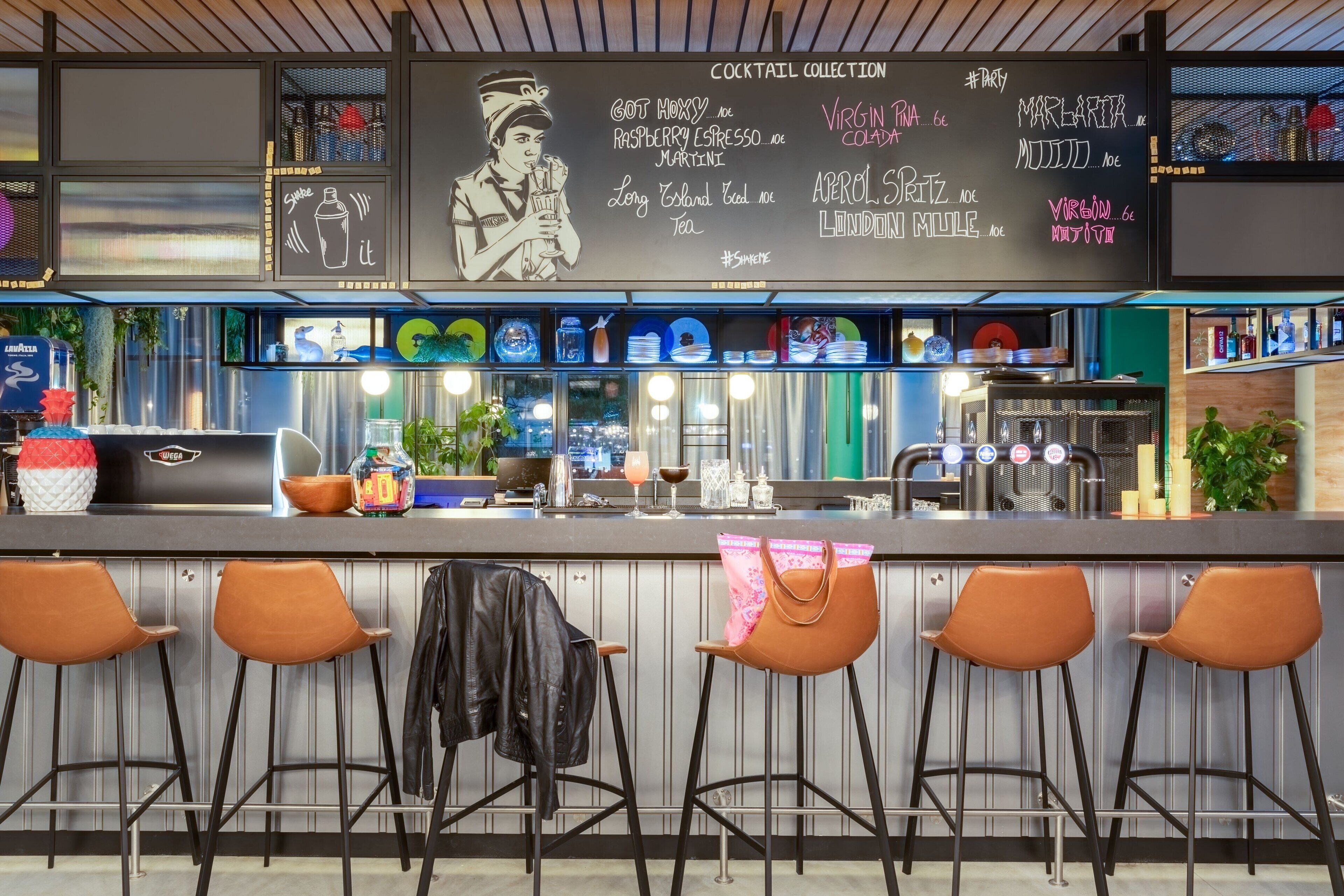 A bar area with a chalkboard menu listing cocktails and a bar counter with stools and various bar tools.