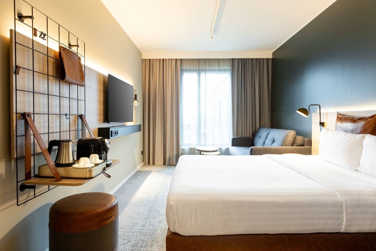 A hotel room with a large bed, a seating area with a sofa and armchairs, a coffee station, a TV mounted on the wall, and a window with curtains.