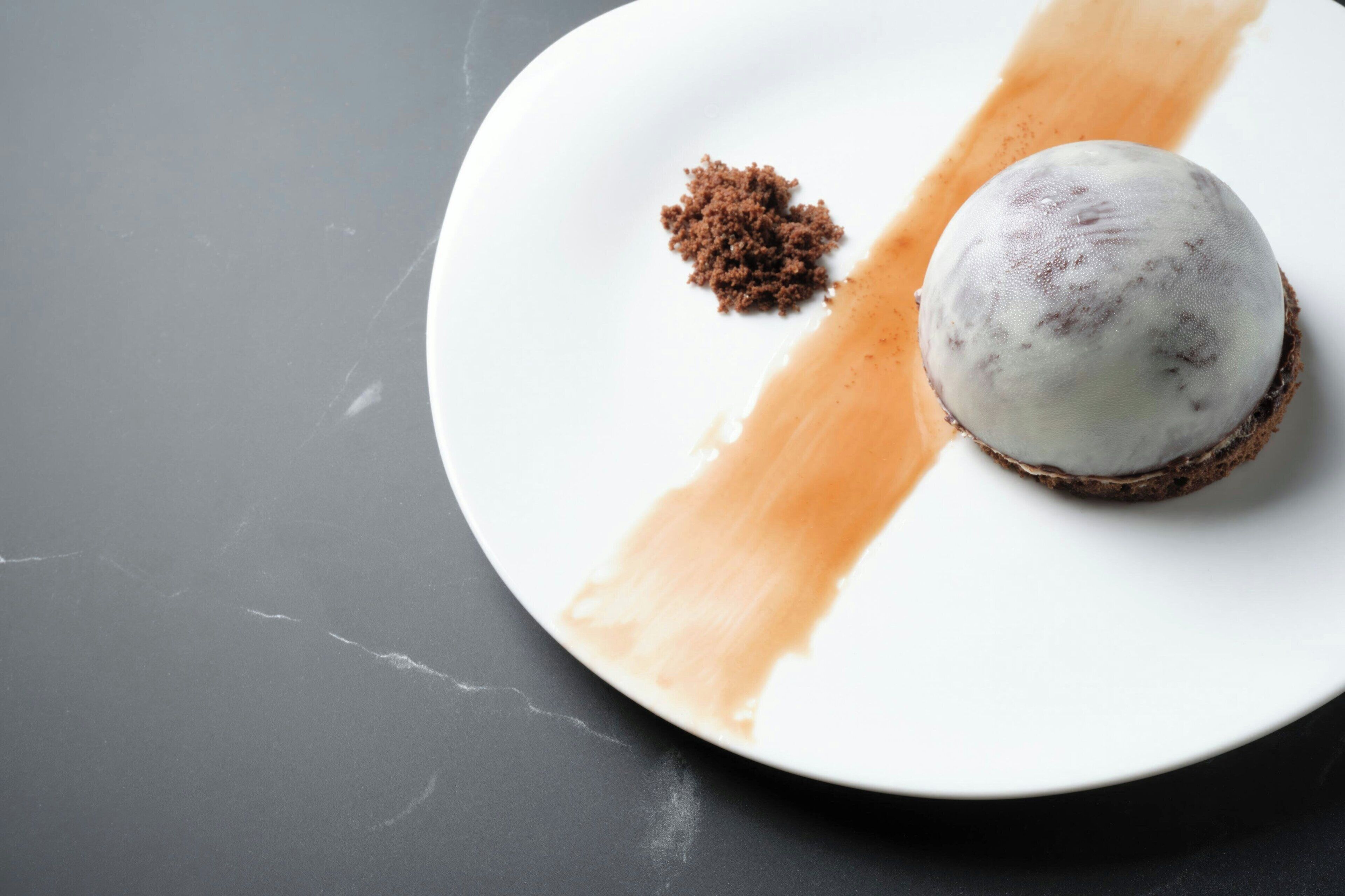 Ein Dessert auf einem weißen Teller mit einem braunen Krumen und einer weißen Kugel, auf einer dunklen Oberfläche platziert.