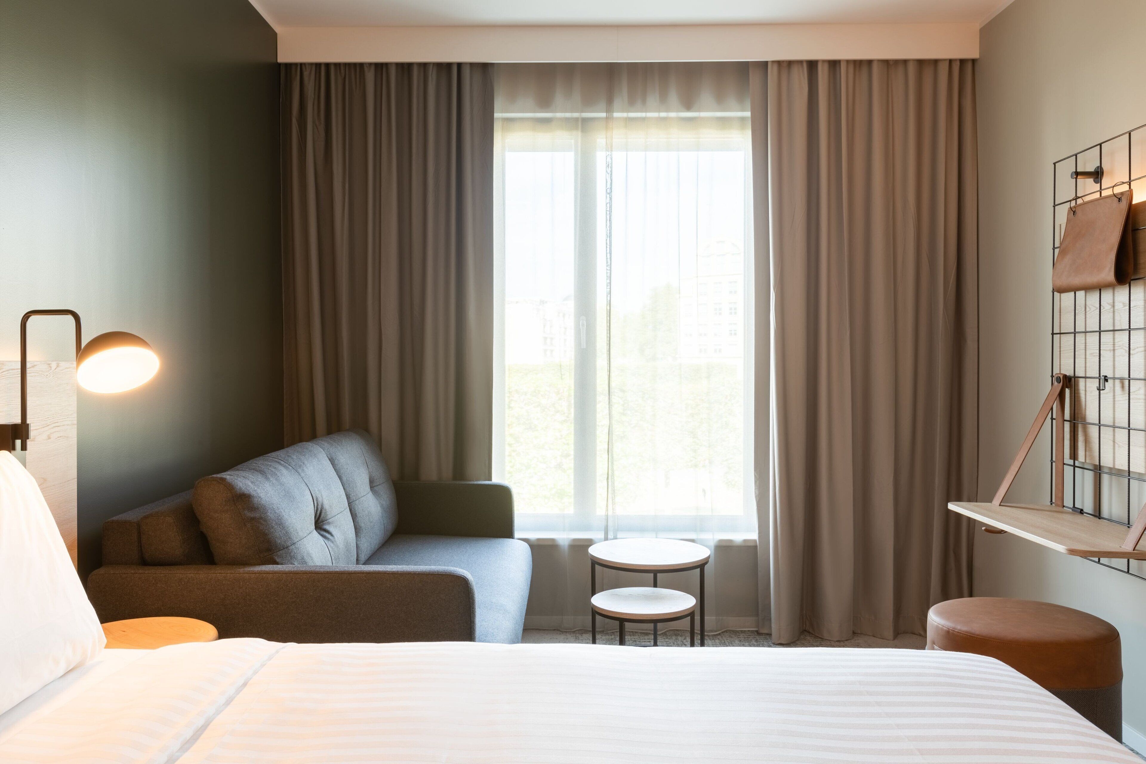 Hotelkamer met een bed, een bank, een klein rond tafeltje en een stoel. Er is een raam met gordijnen, een wandplank met tassen, en een lamp.