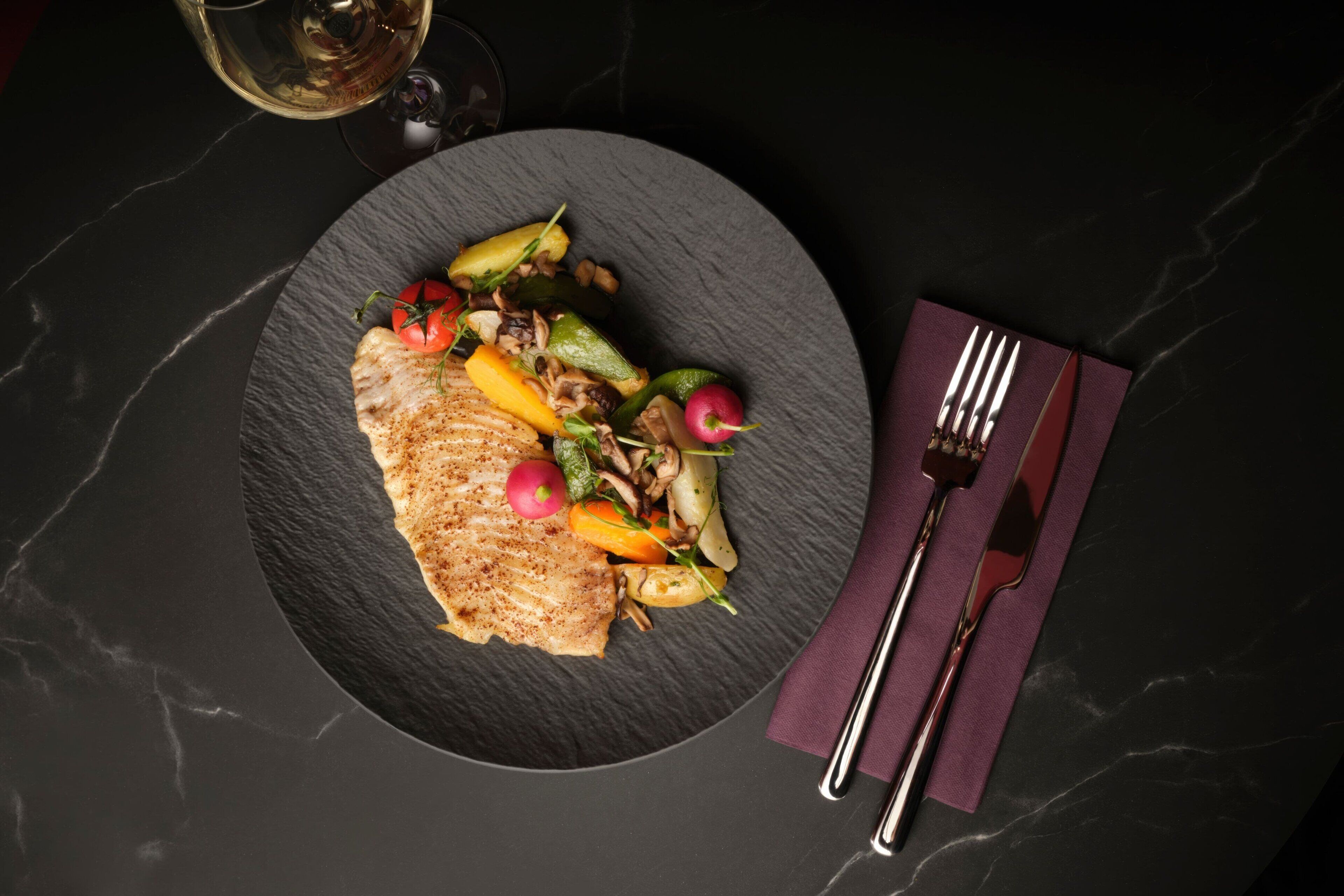 Un plato de comida sobre una mesa de mármol oscuro, con un tenedor y un cuchillo sobre un mantel individual morado.