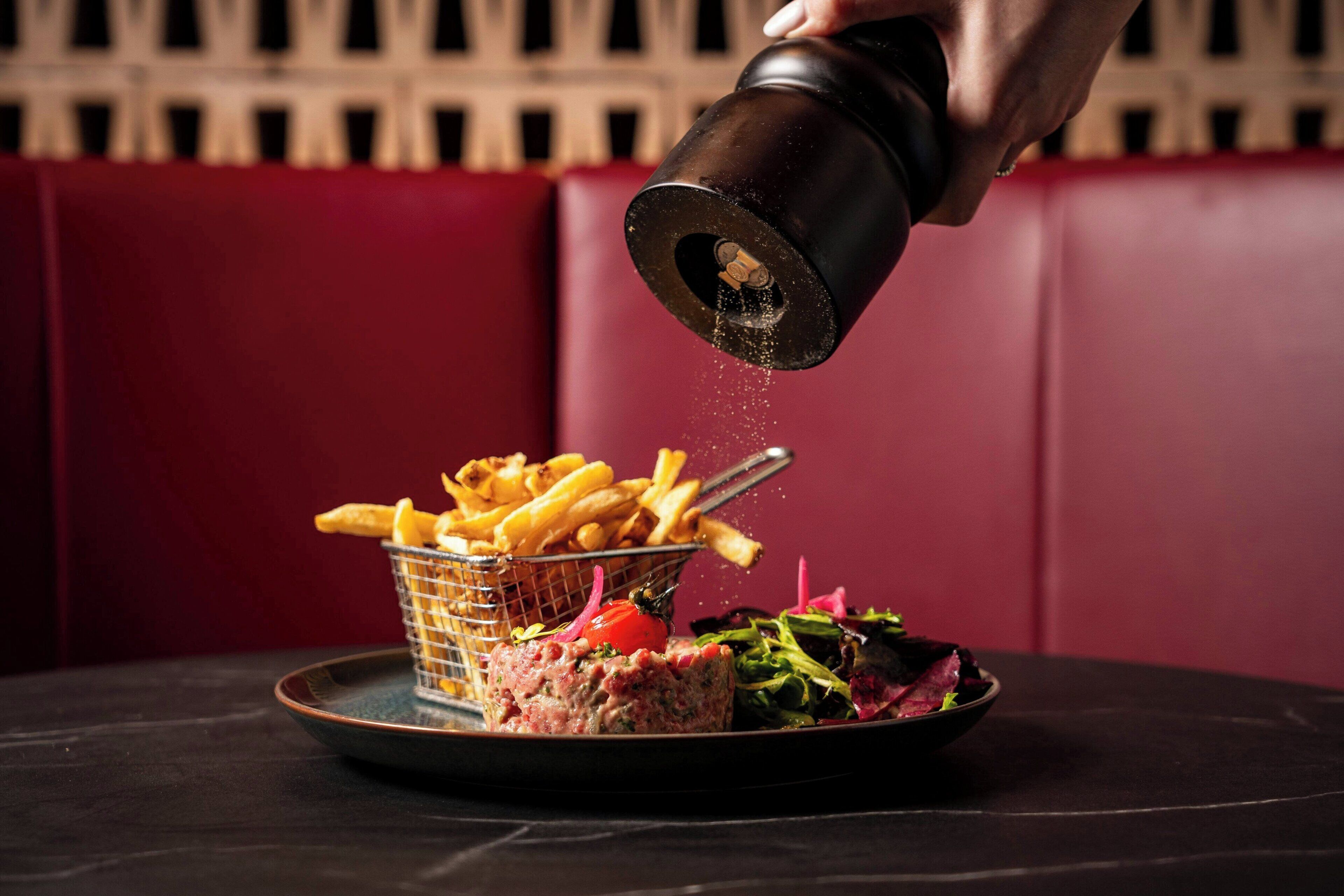 A hand pouring seasoning over a plate of food, including fries and a salad.