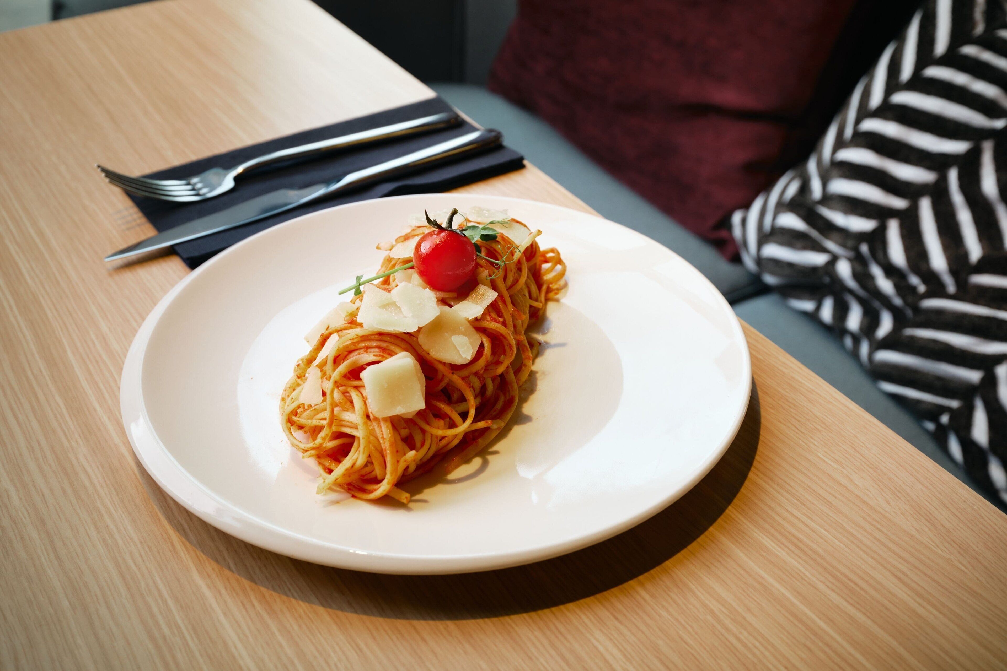Un plato de espagueti con un tomate cherry y queso, sobre un plato blanco, colocado sobre una mesa de madera con un paño de cocina negro y un tenedor al lado.
